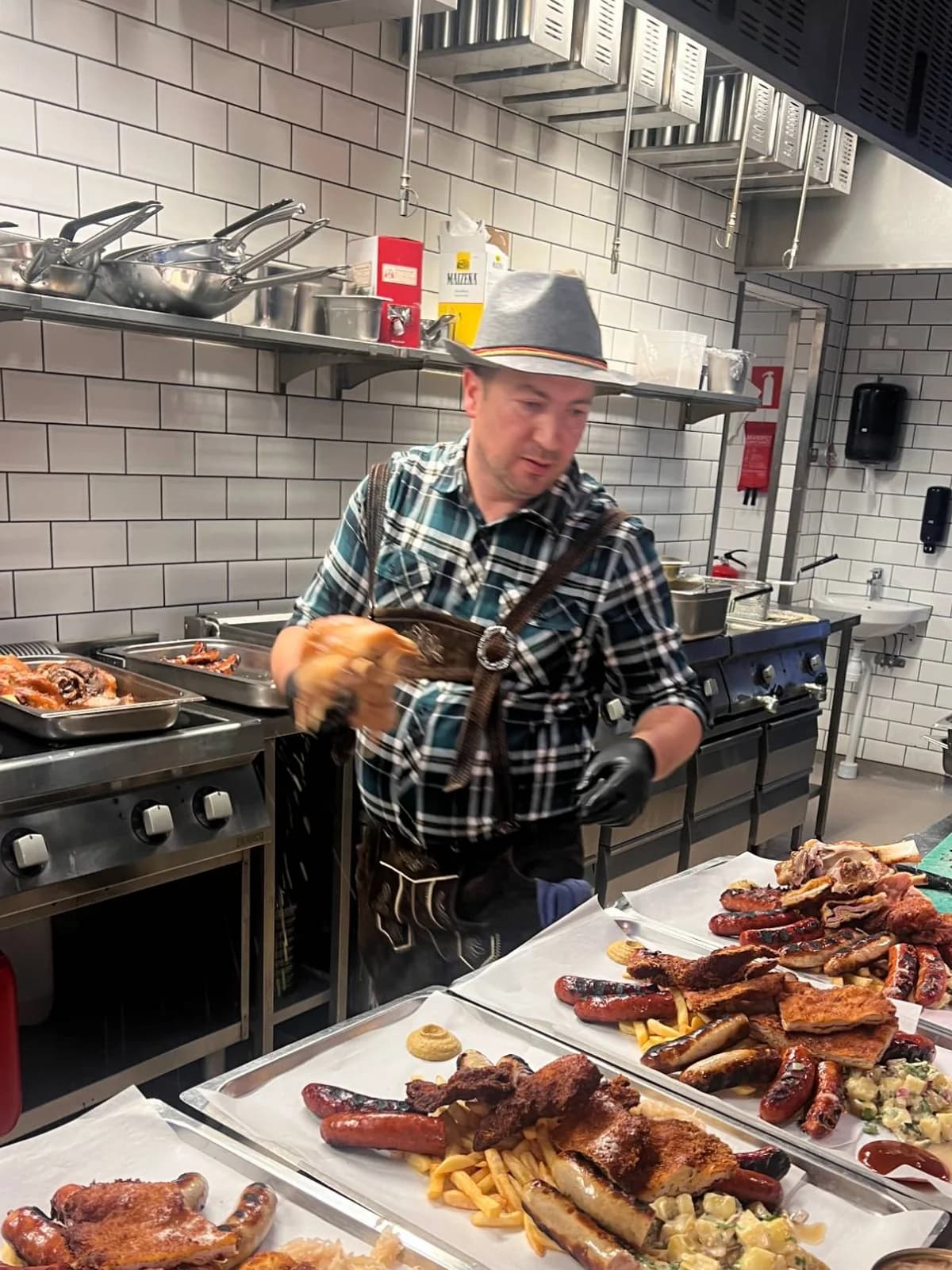 Kock i bayersk hatt och läderbyxor står bakom brickor med grillad korv, schnitzel, pommes och potatissallad i det öppna köket