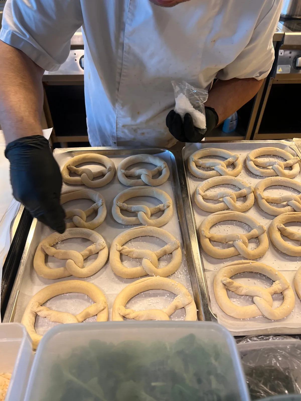Kock i vit rock och svarta handskar strör salt på nyformade kringlor på plåtar i köket