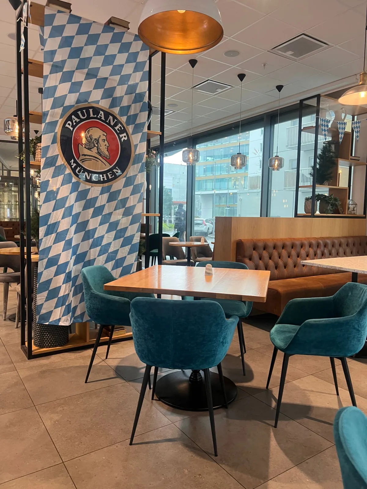Blå-vit Paulaner München-flagga bland turkosa fåtöljer och ett träbord vid fönstret